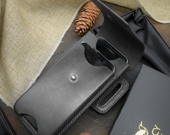 May include: A black leather phone case with a belt loop. The case is open and a smartphone is inside. The case is sitting on a wooden surface.