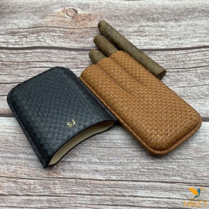 Könnte beinhalten: Zwei Zigarrenetuis aus Leder, eines schwarz und eines braun, mit einem strukturierten Schlangenhautmuster. Das braune Etui enthält drei Zigarren. Das schwarze Etui hat die Initialen "S.J." auf der Vorderseite geprägt.