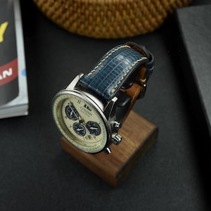 May include: A silver Citizen Eco-Drive watch with a cream face and three sub-dials. It features a blue leather strap with white stitching, displayed on a small wooden stand. The watch face has black numerals and markers.