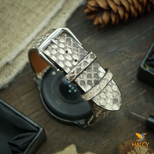 Pode incluir: Uma pulseira de rel&oacute;gio de pele de cobra branca e cinza com uma fivela prateada. A pulseira est&aacute; presa a um smartwatch preto.