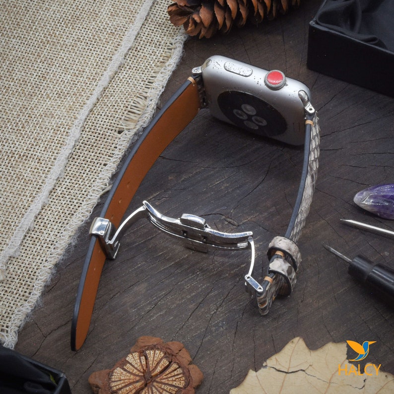 May include: A brown leather Apple Watch band with a snakeskin pattern. The band has a silver buckle and is shown on a wooden surface.