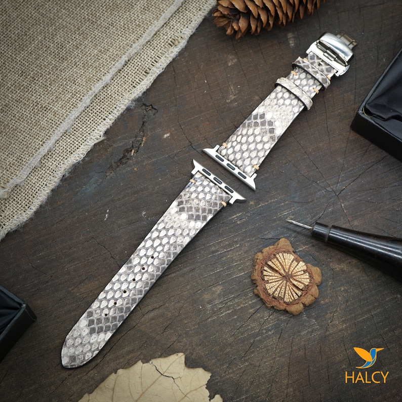 May include: A light gray snakeskin Apple Watch band with silver hardware. The band is laid out flat on a wooden surface.