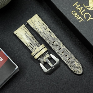 May include: A pair of beige leather watch straps with a textured snake skin pattern. The straps have a silver buckle and are laid out on a black surface.
