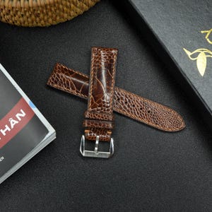 May include: A brown leather watch strap with a textured pattern. The strap has a silver buckle and is made of ostrich leather.