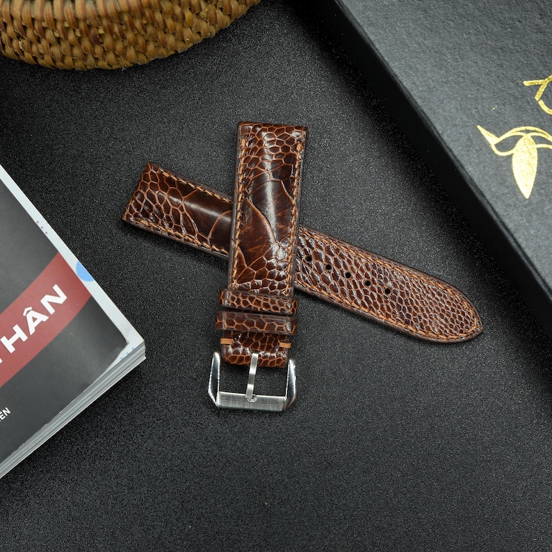 May include: A brown leather watch strap with a textured pattern. The strap has a silver buckle and is made of ostrich leather.