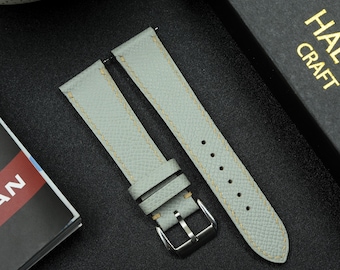 Peut inclure: Un bracelet de montre en cuir bleu clair avec des coutures blanches. Le bracelet est doté d'une boucle et est présenté sur une surface noire.