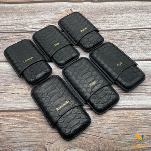 Pode incluir: Uma coleção de estojos de charuto em couro preto com textura de pele de réptil. Cada estojo tem um nome dourado: Leonardo, Eloy, Manuel, Erick, Angel e Raymundo. Os estojos estão dispostos sobre uma superfície de madeira.