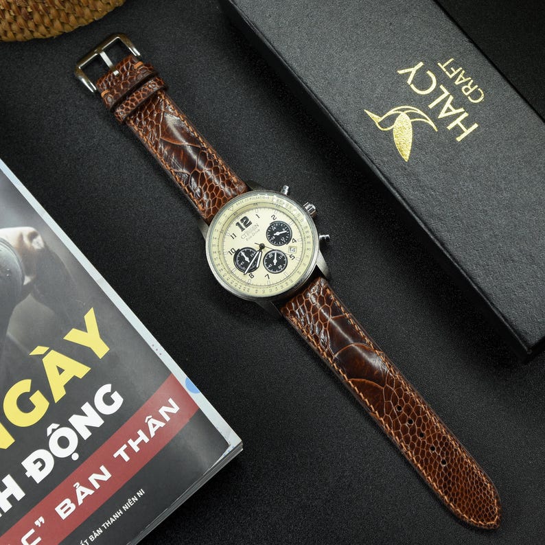 May include: A brown leather watch strap with a textured pattern. The strap is attached to a silver chronograph watch with a white face and black hands. The watch is on a black surface.