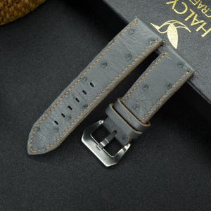 May include: Gray leather watch strap with orange stitching and an ostrich skin pattern. The strap has a silver buckle.
