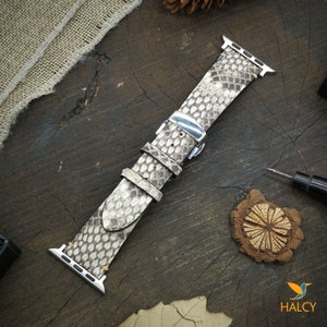 May include: A light gray snakeskin Apple Watch band with a silver buckle. The band is on a wooden surface.