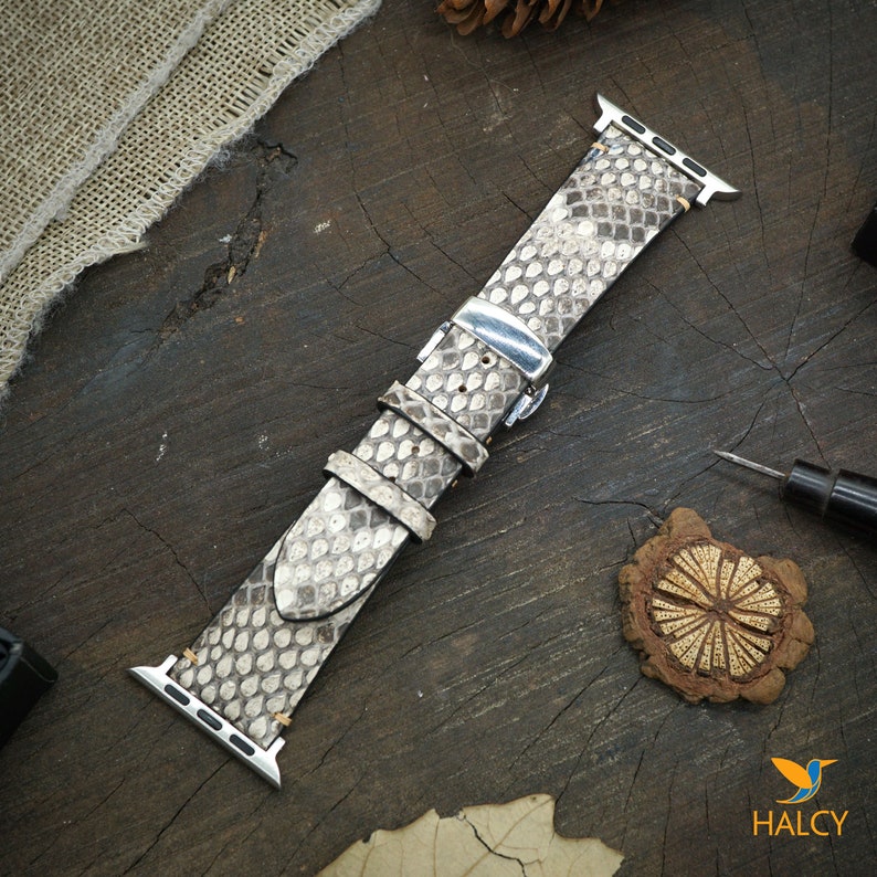 May include: A light gray snakeskin Apple Watch band with a silver buckle. The band is on a wooden surface.