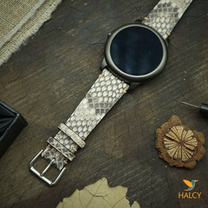 Pode incluir: Uma pulseira de rel&oacute;gio de pele de cobra branca e cinza com uma fivela prateada. A pulseira est&aacute; presa a um smartwatch preto com um mostrador redondo.