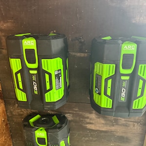 以下が含まれることがあります： 壁に取り付けられた黒と緑のEgo 56V ARCリチウムバッテリー3個。バッテリーには、Egoのロゴと「56V」と「ARCリチウム」の文字が書かれています。