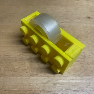 May include: A yellow plastic tape dispenser shaped like a Lego brick. The dispenser holds a roll of clear tape.