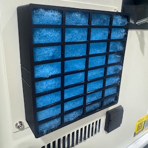 May include: A close-up of a black air filter with a blue filter inside. The filter is mounted on a white surface.