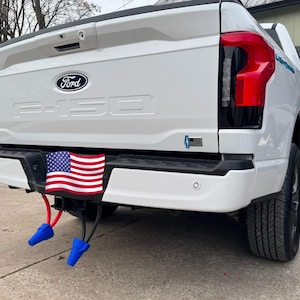 May include: The back of a white Ford pickup truck with a US flag hanging from the hitch. The truck has a black bumper and black taillights. There are two blue electrical connectors hanging from the hitch.