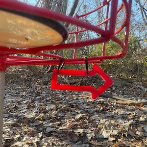 May include: A red disc golf basket with a red arrow pointing to the right, hanging from the top of the basket. The basket is in a wooded area with fallen leaves on the ground.