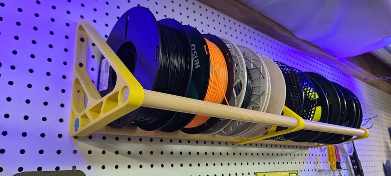 Op de afbeelding: Een wandbevestigde spoelhouder met een houten pen en gele kunststof beugels. De houder is gevuld met spoelen filament, waaronder zwart, oranje en grijs.