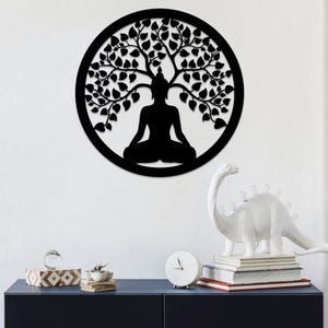 May include: A black, circular wall art piece featuring a silhouette of a seated Buddha figure under a tree with leafy branches. The artwork is set against a white wall, with decorative items on a dark blue surface below.