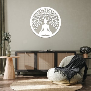 May include: A white, circular wall art piece featuring a meditating figure beneath a tree. The design is cut out, creating a silhouette effect. The artwork is mounted on a neutral-colored wall, adding a touch of serenity to the space.