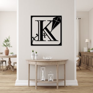 May include: Black metal wall art featuring the letter K with a kite design. The art is framed in a square and is mounted on a white wall above a wooden console table.