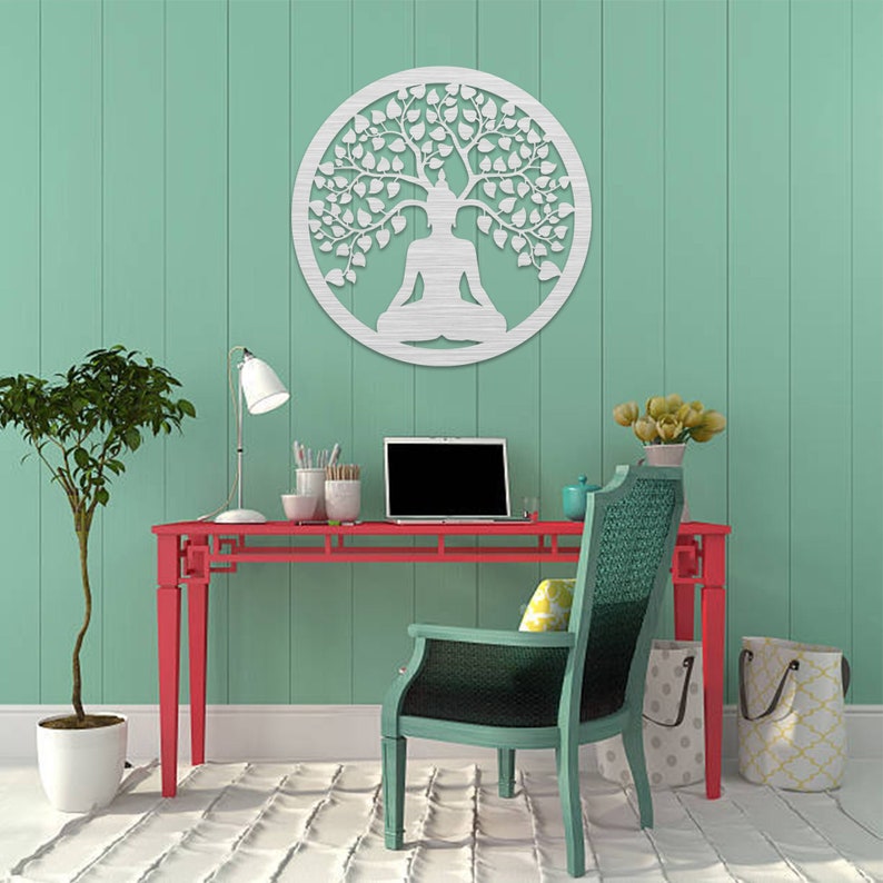 May include: A round, silver-colored metal wall art piece depicting a meditating figure under a tree. The artwork is mounted on a light green wall above a red desk. A laptop, lamp, and chair are also visible.