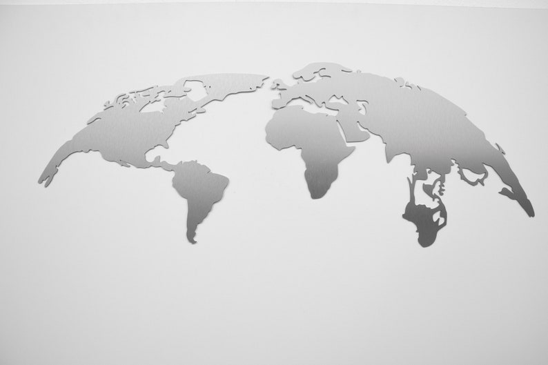 May include: A silver-colored metal world map wall art. The map is a detailed outline of the continents, including North and South America, Europe, Africa, Asia, and Australia. The artwork is mounted on a white wall.