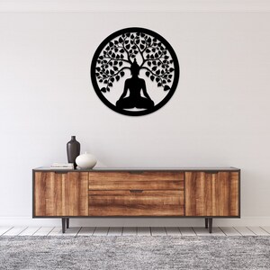 May include: Black metal wall art depicting a meditating Buddha under a tree, enclosed in a circular frame. The artwork is mounted on a white wall above a wooden cabinet with a gray rug.