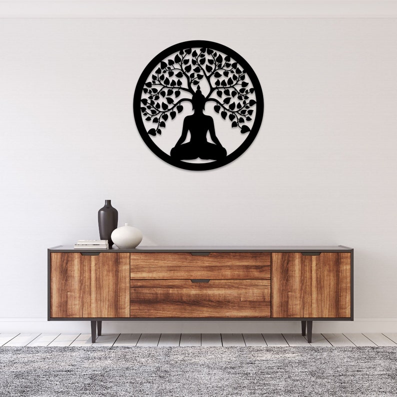 May include: Black metal wall art depicting a meditating Buddha under a tree, enclosed in a circular frame. The artwork is mounted on a white wall above a wooden cabinet with a gray rug.