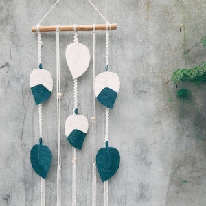 Może przedstawiać: Makrama ścienna z motywami w kształcie liści w kolorze białym i turkusowym, zawieszona na naturalnym drewnianym kołku. Liście ułożone są pionowo, z drewnianymi koralikami dodającymi szczegółów. Tło to teksturowana szara ściana.