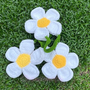 May include: Three crocheted white daisy flowers with yellow centers. The flowers are arranged in a cluster on a green grassy surface.