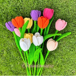 May include: A bouquet of handmade crochet tulips in various colors, including orange, purple, pink, yellow, and white. The flowers have green stems and leaves, arranged on a bed of green grass. The overall theme is spring and handmade crafts.