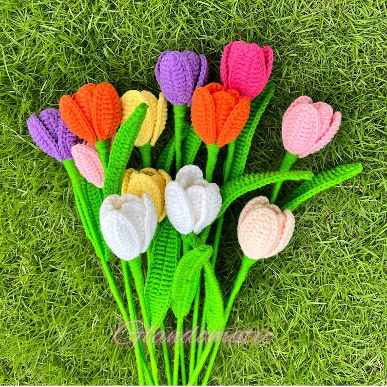 May include: A bouquet of handmade crochet tulips in various colors, including orange, purple, pink, yellow, and white. The flowers have green stems and leaves, arranged on a bed of green grass. The overall theme is spring and handmade crafts.