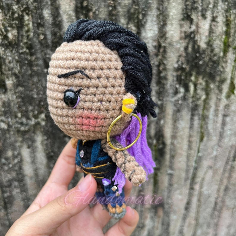 K&ouml;nnte beinhalten: H&auml;kelpuppe Amigurumi mit schwarzen Haaren, lila Z&ouml;pfen und goldenen Ohrringen. Die Puppe tr&auml;gt ein blaues und schwarzes Outfit mit lila Akzenten.