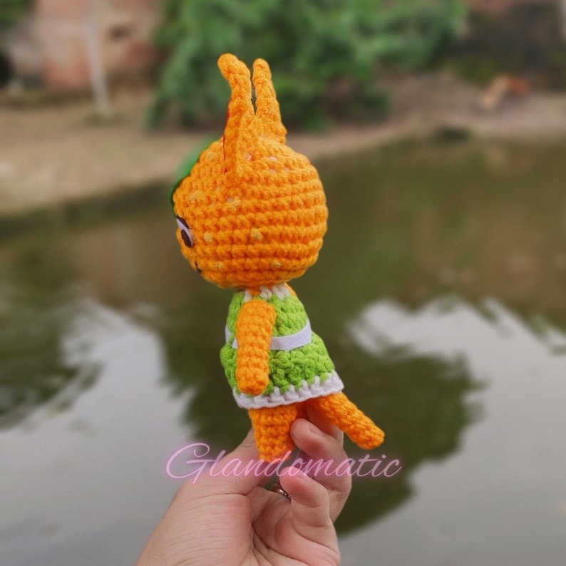 Pu&ograve; includere: Peluche di animali in crochet arancione e verde con un vestito bianco. Il giocattolo &egrave; tenuto in una mano.