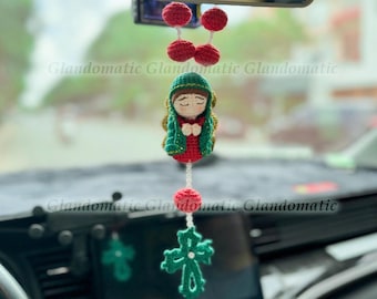 May include: Crocheted car charm featuring a religious figure with a green and red outfit, a green veil, and a cross pendant. The charm includes red crocheted beads and is suspended from a string.