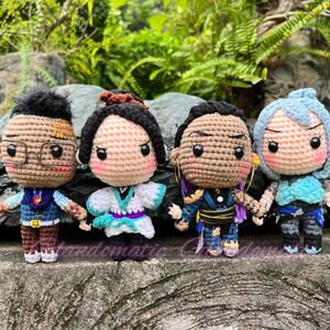 K&ouml;nnte beinhalten: Vier geh&auml;kelte Amigurumi-Puppen mit verschiedenen Gesichtsausdr&uuml;cken und Outfits. Die Puppen tragen blaue, gr&uuml;ne und violette Kleidung. Die Puppen stehen auf einer braunen Oberfl&auml;che.