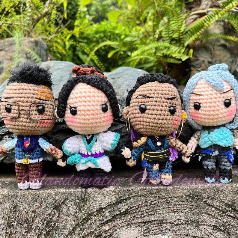 K&ouml;nnte beinhalten: Vier geh&auml;kelte Amigurumi-Puppen mit verschiedenen Gesichtsausdr&uuml;cken und Outfits. Die Puppen tragen blaue, gr&uuml;ne und violette Kleidung. Die Puppen stehen auf einer braunen Oberfl&auml;che.