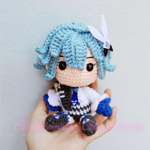 May include: A crocheted plush toy with blue hair, a white shirt, and black and white pants. The toy has a white bunny ear headband with the text "73" on it.