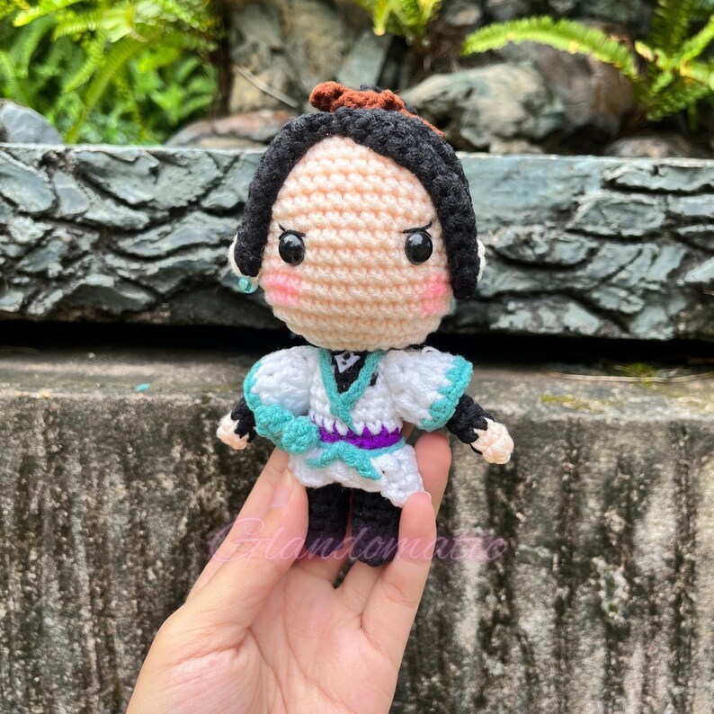 Pu&ograve; includere: Bambola di un personaggio anime realizzata a uncinetto con capelli neri, un vestito bianco e blu e una fascia viola. La bambola ha occhi grandi e un blush rosa.
