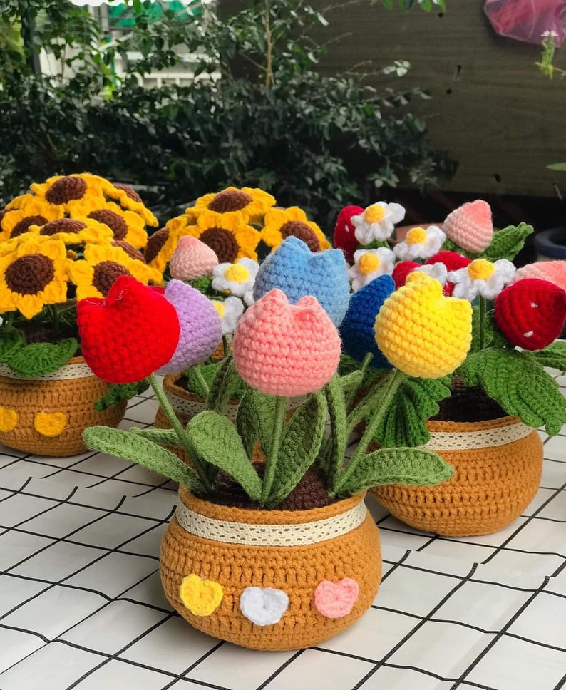 Peut inclure: Un ensemble de pots de fleurs en crochet avec des fleurs color&eacute;es. Les pots sont bruns avec une bordure en dentelle blanche et des c&oelig;urs jaunes, blancs et roses. Les fleurs comprennent des tournesols, des marguerites et des tulipes dans des tons de rouge, rose, bleu, jaune et violet.