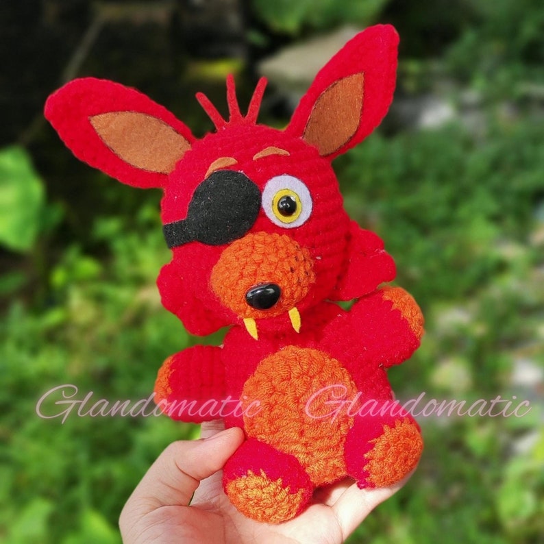 Puede incluir: Un juguete de peluche rojo y naranja tejido a crochet con un parche en el ojo negro y un ojo amarillo. El juguete tiene una sonrisa de dientes y tiene los brazos extendidos.
