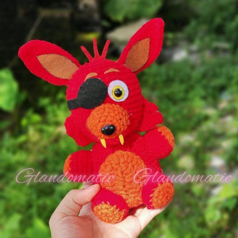 Puede incluir: Un juguete de peluche rojo tejido a crochet con una barriga naranja y un parche en el ojo negro. El juguete tiene un ojo blanco y una nariz negra. El juguete est&aacute; siendo sostenido en una mano.
