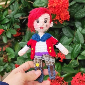 May include: A crocheted doll with red hair, a red jacket, a blue shirt, and a purple belt. The doll is wearing brown boots and has a yellow and white design on its chest. The doll is holding a small, white object in its hand.