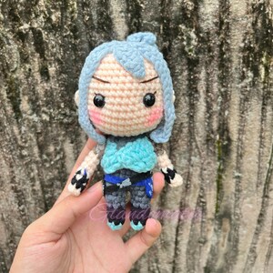 Pu&ograve; includere: Una bambola all'uncinetto con capelli blu, un top blu e bianco e pantaloni grigi. La bambola ha un viso rosa chiaro con occhi neri e guance rosa.