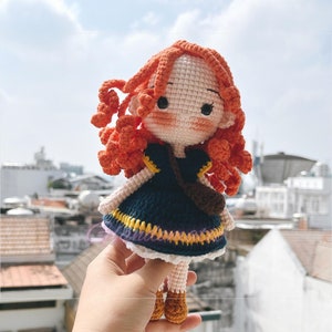May include: Crocheted doll with orange curly hair, wearing a navy blue dress with yellow and white trim, and a brown shoulder bag. The doll has a light skin tone and is holding a small brown bag. The doll is handmade.