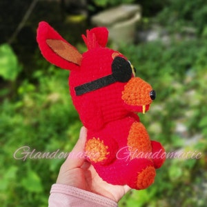 Puede incluir: Juguete de peluche rojo de crochet con un parche en el ojo, que se asemeja a un animal de dibujos animados con orejas grandes y una amplia sonrisa.
