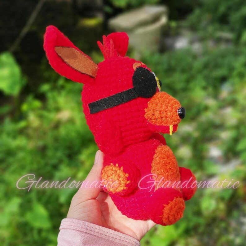 Puede incluir: Juguete de peluche rojo de crochet con un parche en el ojo, que se asemeja a un animal de dibujos animados con orejas grandes y una amplia sonrisa.