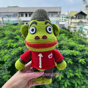 May include: A handmade crochet doll in the shape of a cartoon monkey. The monkey has a green face, ears, and feet, with a red mouth and a dark green cap. It wears a red shirt with a white number 1 and a white patch. The doll is held in front of a green leafy background.