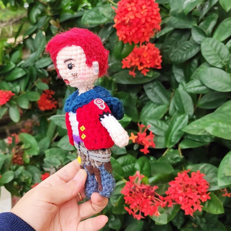 May include: A crocheted doll with red hair, a blue and red jacket, and blue pants. The doll is holding a red and white heart in its hand. The doll is standing in front of a green bush with red flowers.